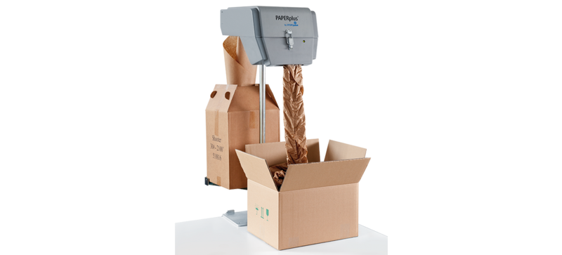 A cardboard box on a table, a gray machine feeding brown paper cushioning into the box